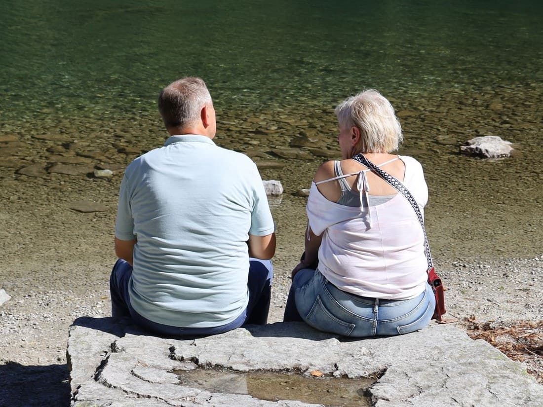 An older couple sitting together on the shore of a mountain lake, looking out over the water
