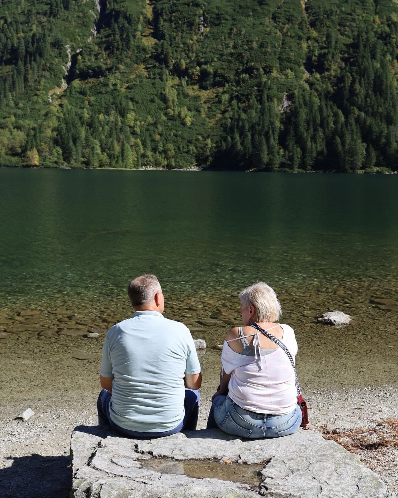 An older couple sitting together on the shore of a mountain lake, looking out over the water